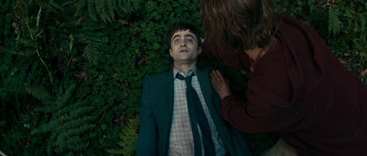 Un pallido uomo ferito (Daniel Radcliffe) in abito azzurro e cravatta sdraiato a faccia in su in un campo di fogliame esotico mentre un uomo dai capelli lunghi con una felpa bordeaux (Paul Dano) si china su di lui.
