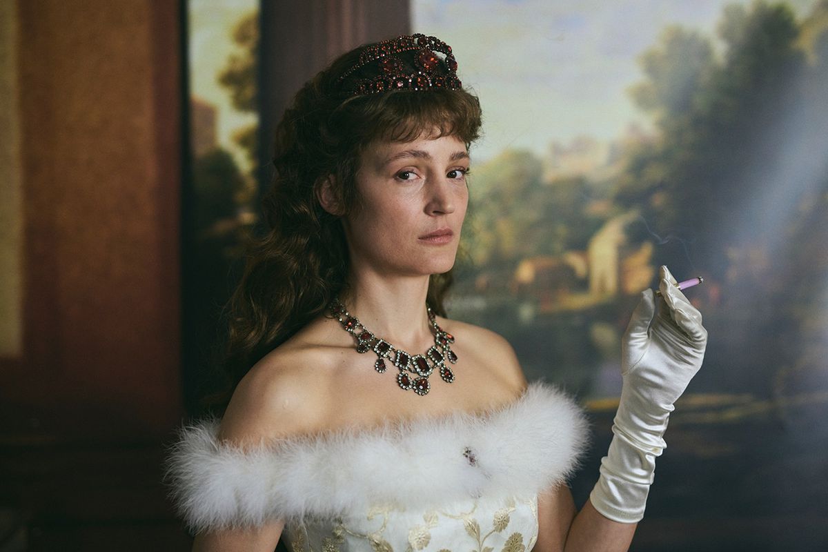 Vicky Krieps fuma una sigaretta e indossa una corona, un costume e un guanto bianco davanti a un dipinto ornato nei panni dell'imperatrice Elisabetta d'Austria in Corsage.