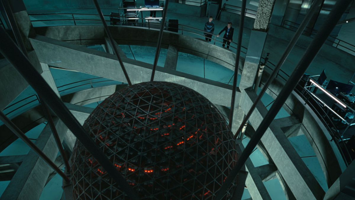 un uomo e una donna guardano oltre il bordo di un edificio circolare per vedere Rehoboam, un'IA sferica che brilla di rosso