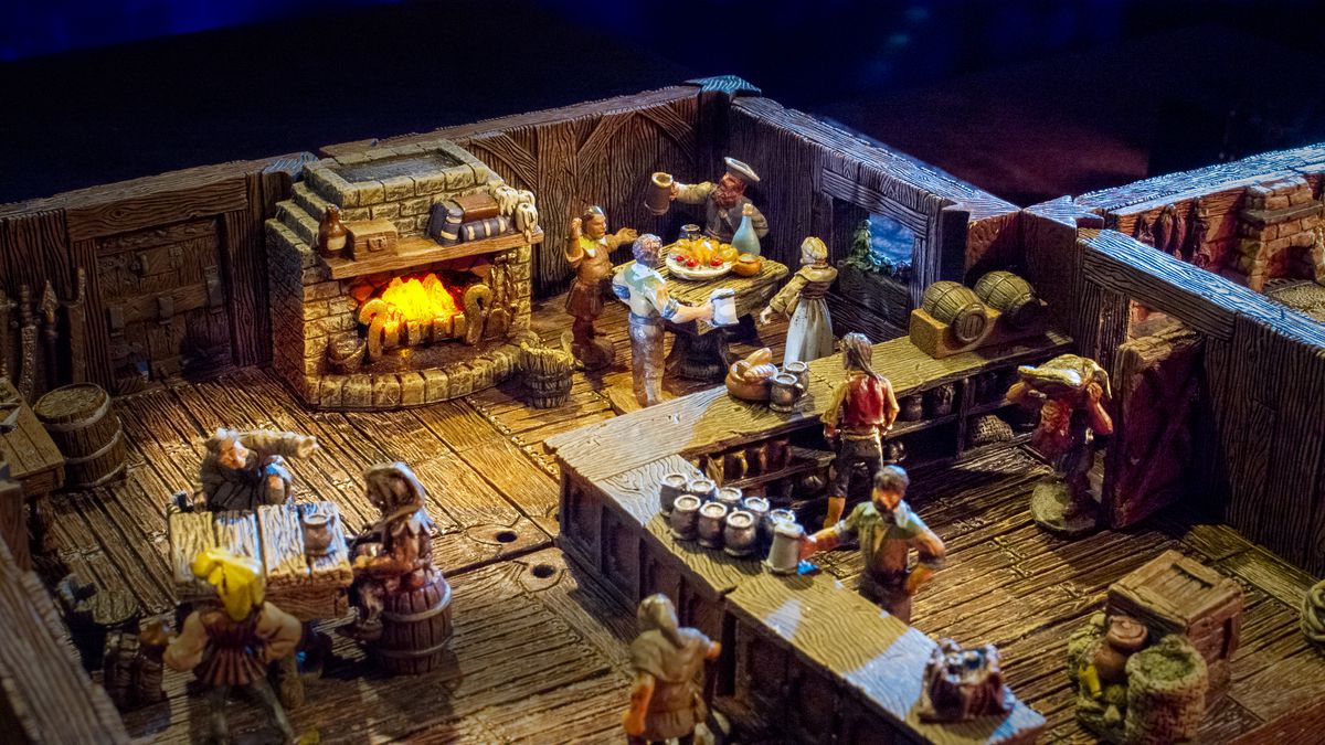 Un'inquadratura ampia di un gruppo di avventori in una taverna medievale.  Le birre sono accatastate sul bancone e un pasto viene condiviso nell'angolo, tutto in miniatura, utilizzando il terreno Dwarven Forge.