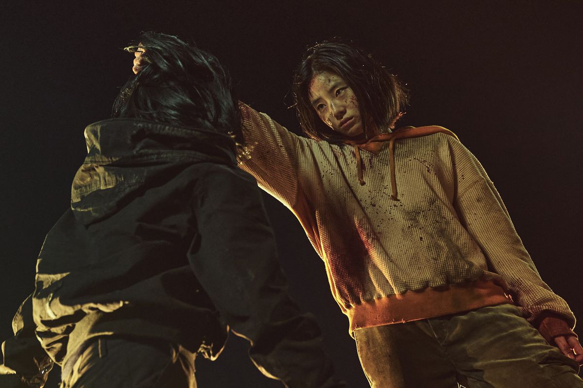 Una giovane ragazza (Shin Si-ah) con una felpa arancione insanguinata tiene un uomo con una giacca nera per i capelli.