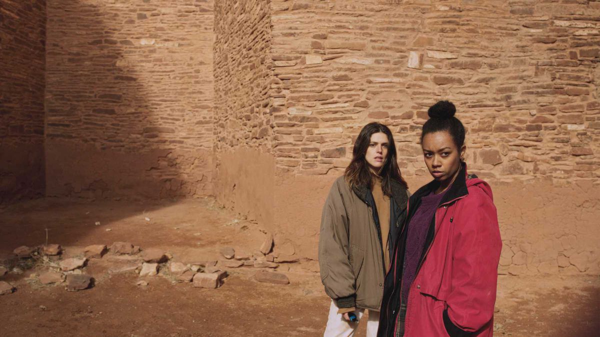 Due giovani donne si trovano di fronte a una struttura in mattoni di argilla e guardano in lontananza a Jethica.