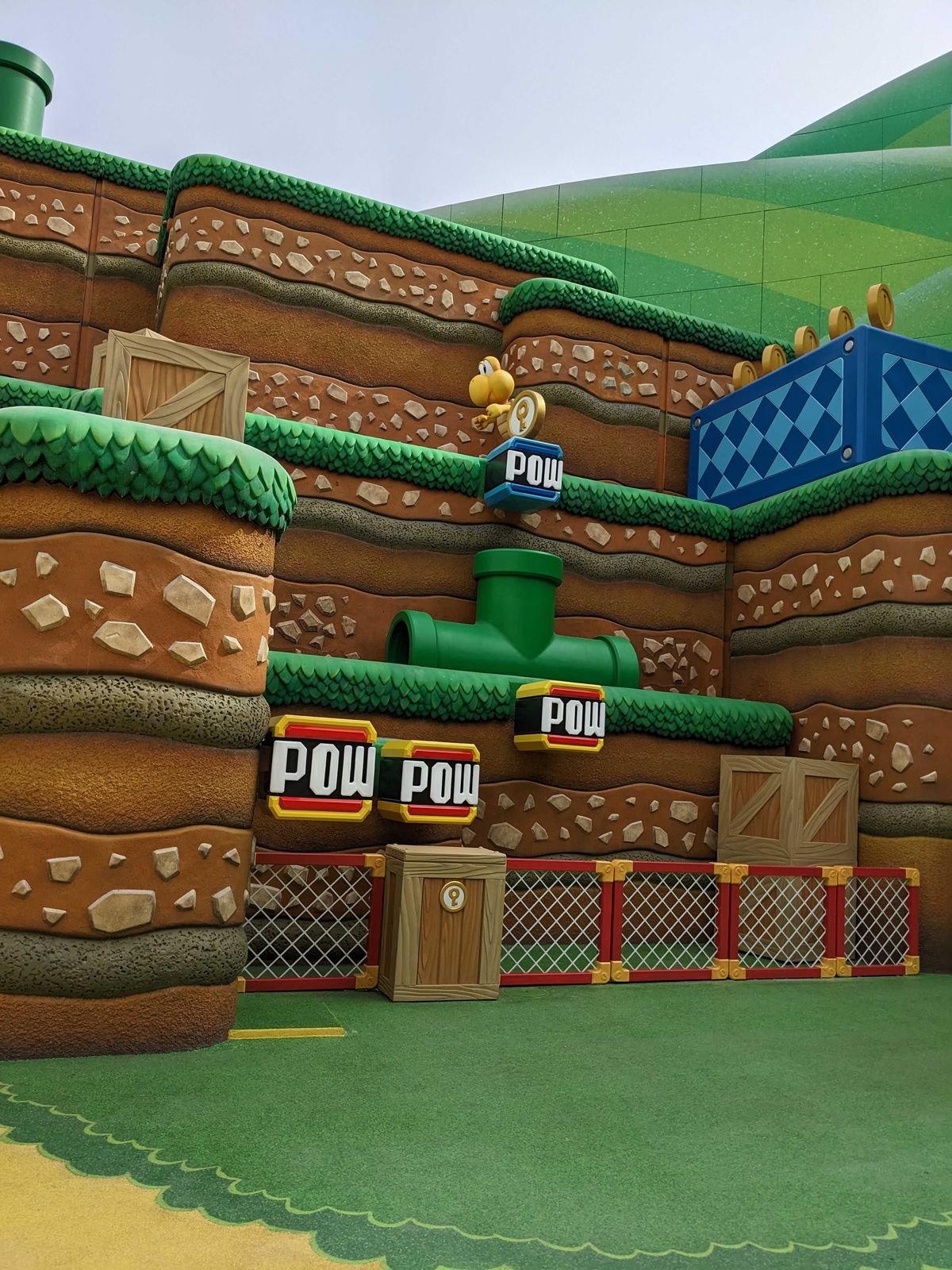 Un Koopa si trova sopra un pulsante POW, posizionato sopra un tubo.  L'attività fa parte di Super Nintendo World.