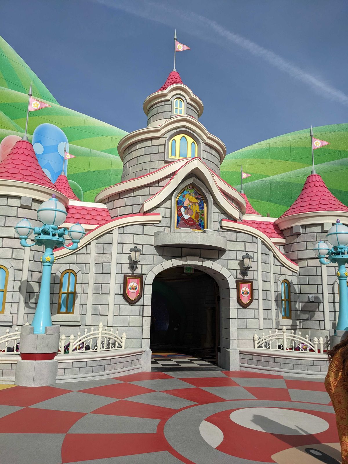 Il castello della Principessa Peach in Super Nintendo World.  Il castello è grigio, ha più piani, tetto rosa, un'immagine in vetro colorato di una Principessa Peach e una bandiera in cima.