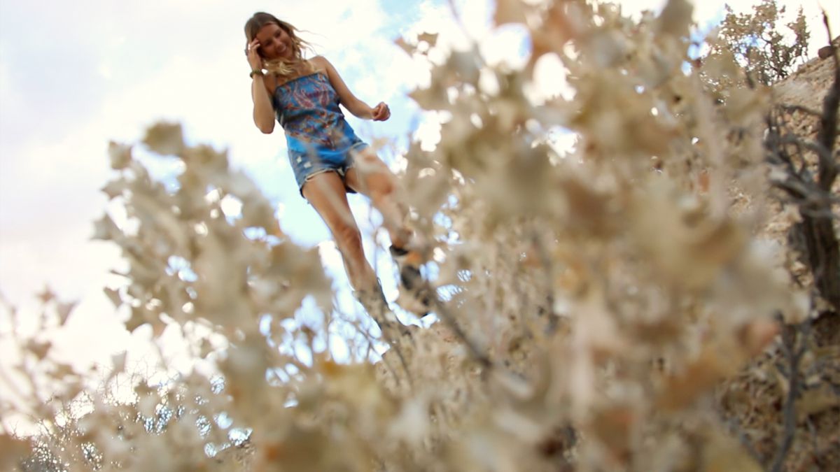 Una giovane donna bionda che indossa un top colorato è vista attraverso le piante del deserto nelle Outwaters.
