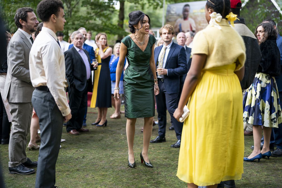Una giovane donna di colore con un vestito giallo brillante si trova in mezzo a una folla di persone all'aperto in quella che sembra essere una festa in giardino e urla contro di loro in una scena di The Strays di Netflix