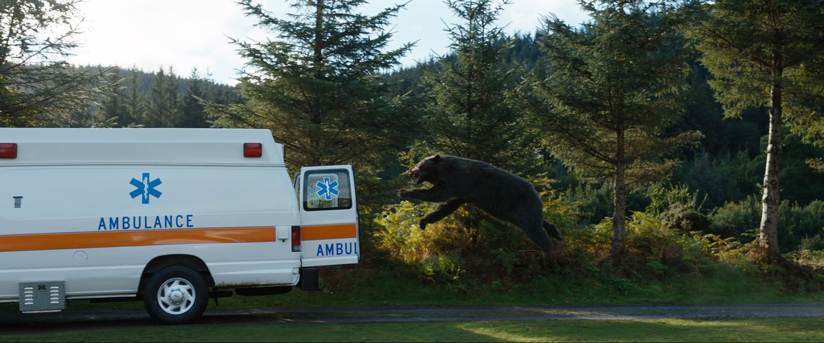 Un orso CGI fa un salto in volo nel retro scoperto di un'ambulanza, visto in una ripresa d'azione a lunga distanza, in Cocaine Bear