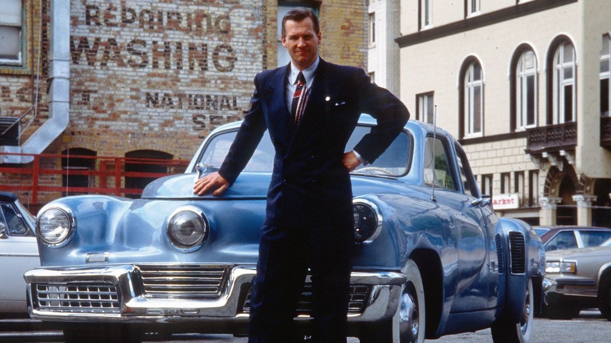Un uomo con i capelli pettinati all'indietro in un abito blu scuro (Jeff Bridges) è in piedi davanti a un'automobile degli anni '40.
