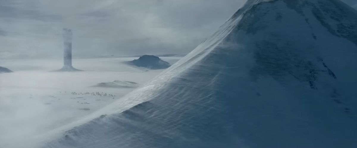 Una guglia nera spettrale parzialmente nascosta dalle nuvole.  Una montagna si staglia in primo piano, ricoperta di neve.
