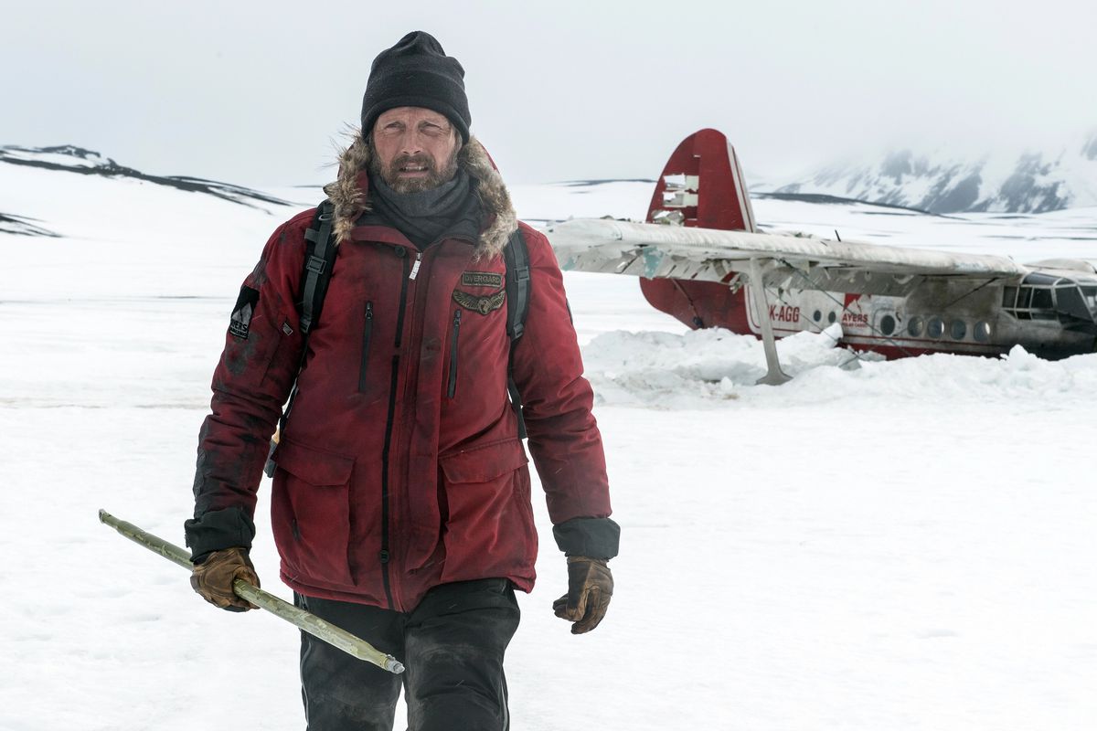 Un uomo con una giacca invernale rossa (Mads Mikkelsen) si allontana da un aereo abbattuto circondato da neve e montagne.