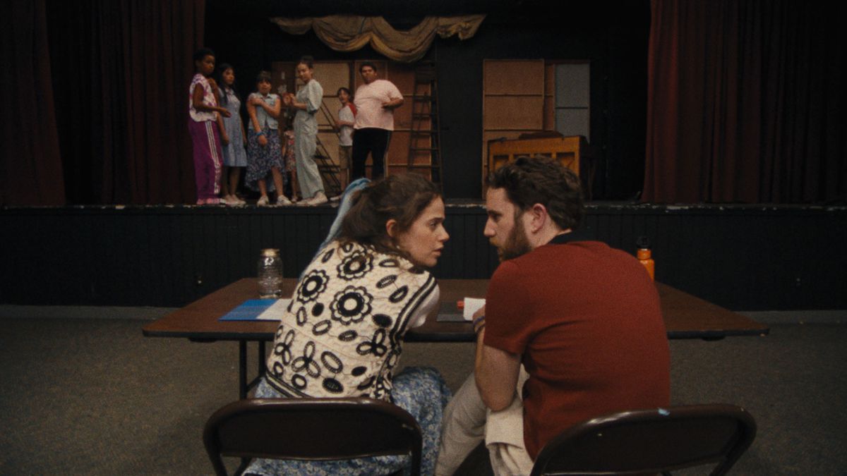Molly Gordon e Ben Platt parlano tra loro dietro un tavolo in Theatre Camp, mentre gli attori sul palco guardano.