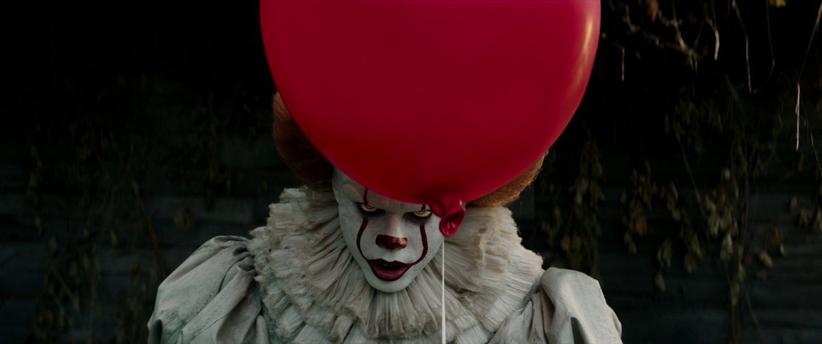Un clown con la pittura facciale bianca e rossa guarda da dietro un palloncino rosso vivo che galleggia davanti al loro viso.