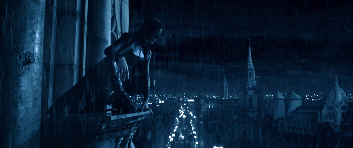 Una donna dai capelli corti con una tuta di pelle e un cappotto nero è inginocchiata sul bordo di un edificio che domina una città di notte con i campanili delle chiese e gli edifici in lontananza.