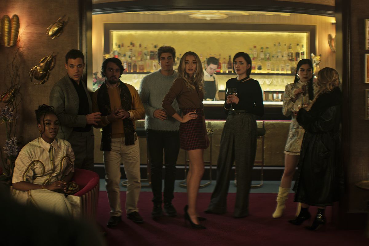 Un piccolo gruppo di persone molto ricche si trova di fronte a un bar e posa per la telecamera nella quarta stagione di You di Netflix
