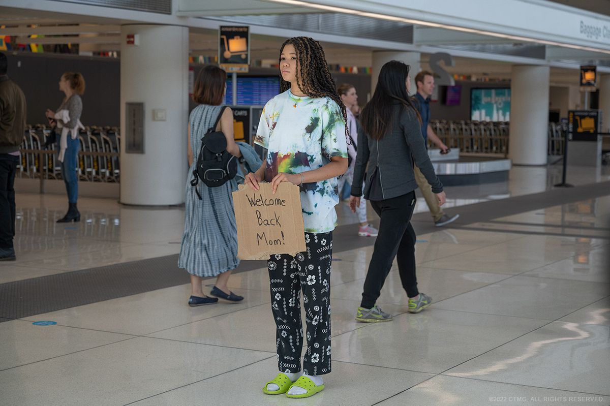 Storm Reid, che indossa il pigiama + crocs fit che si indossa in aeroporto, tiene in mano un cartello di cartone che dice 