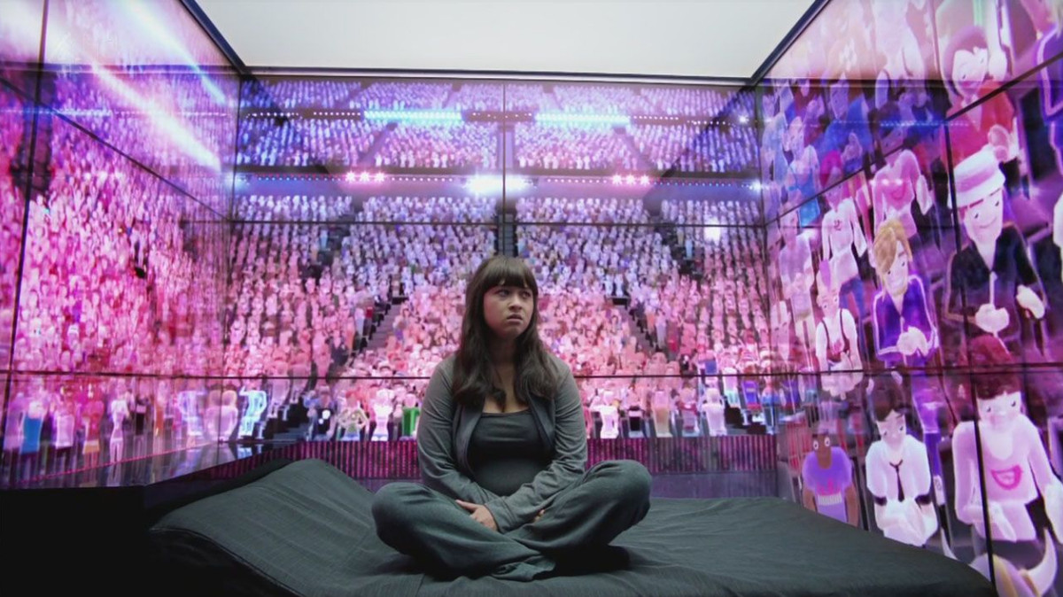 Una donna siede in una stanza circondata da pareti di avatar di computer seduti in un grande auditorium.