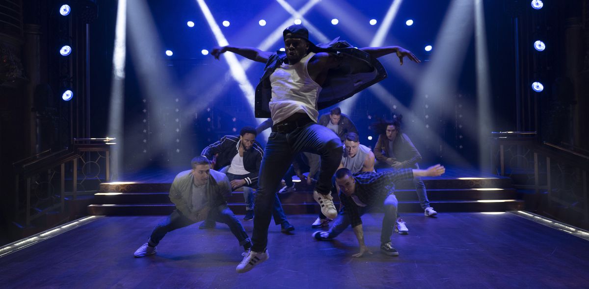 Un gruppo di ballerini maschi (completamente vestiti, mi dispiace) accovacciati su un palco illuminato da una luce blu intenso e tratteggiato da riflettori mentre un ballerino salta verso l'alto con le braccia aperte in Magic Mike's Last Dance