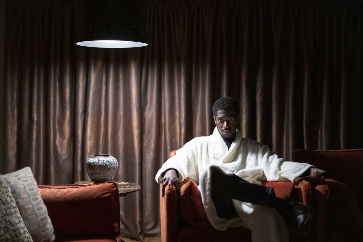 Un giovane uomo di colore (Jorden Myrie) in un soffice accappatoio bianco siede su una lussuosa sedia rossa in una stanza buia con tende e fissa direttamente la telecamera in una scena di The Strays di Netflix