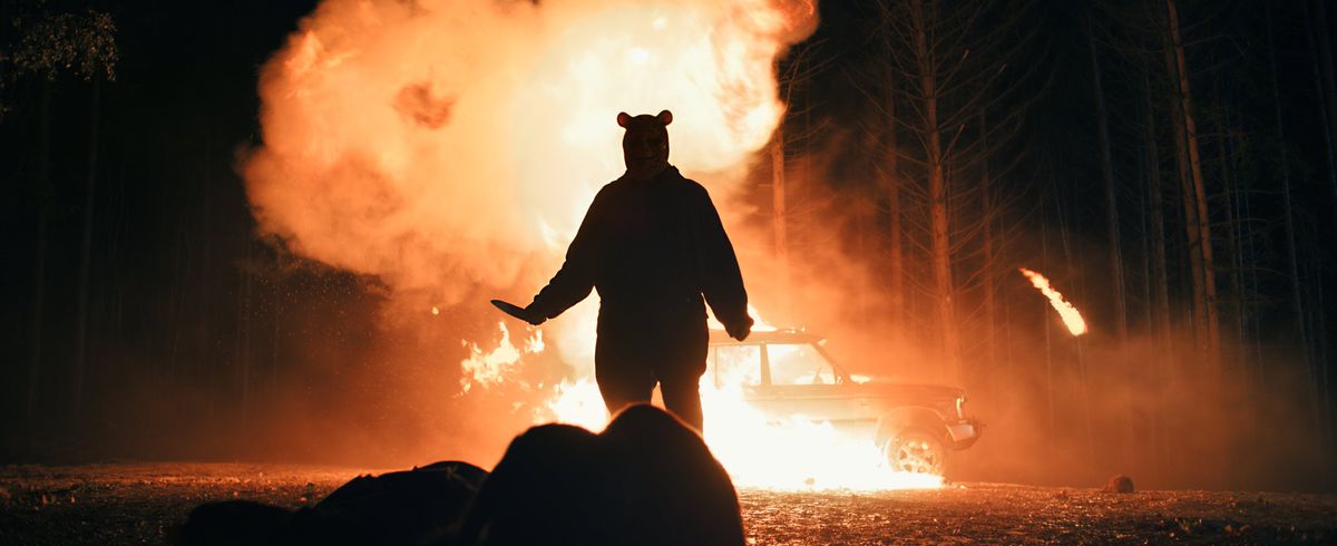 Winnie the Pooh (Craig David Dowsett) si staglia contro un camion che esplode nel bosco di notte e alza un coltello mentre si avvicina a un'altra vittima in Winnie the Pooh: Blood and Honey