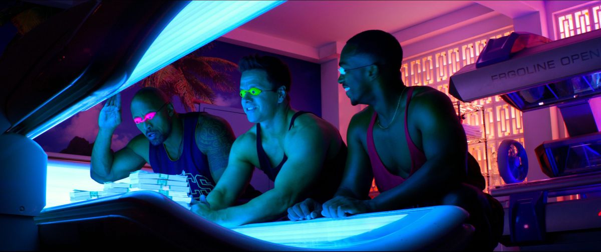 Dwayne Johnson, Mark Wahlberg e Anthony Mackie sorridono davanti a un lettino abbronzante mentre indossano canotte colorate in Pain and Gain.