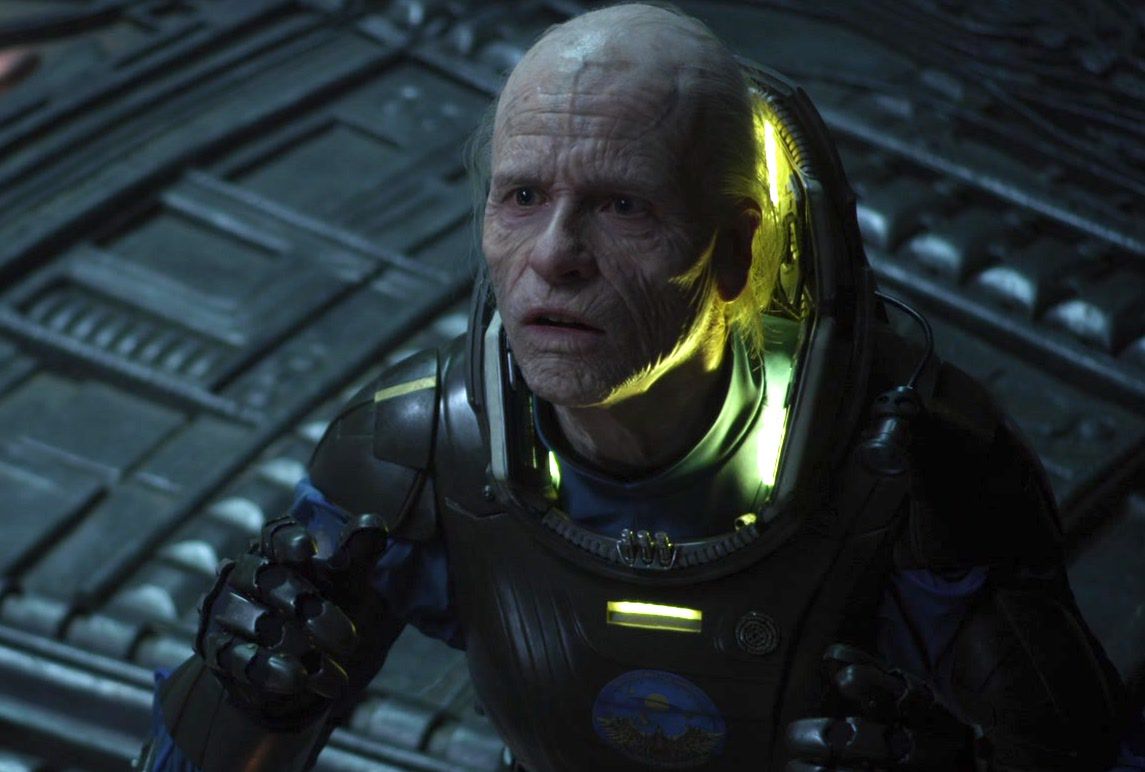 Il vecchio Weyland (Guy Pearce con il trucco per la vecchiaia) in una tuta spaziale si inchina a una presenza fuori campo in un'astronave aliena in Prometheus