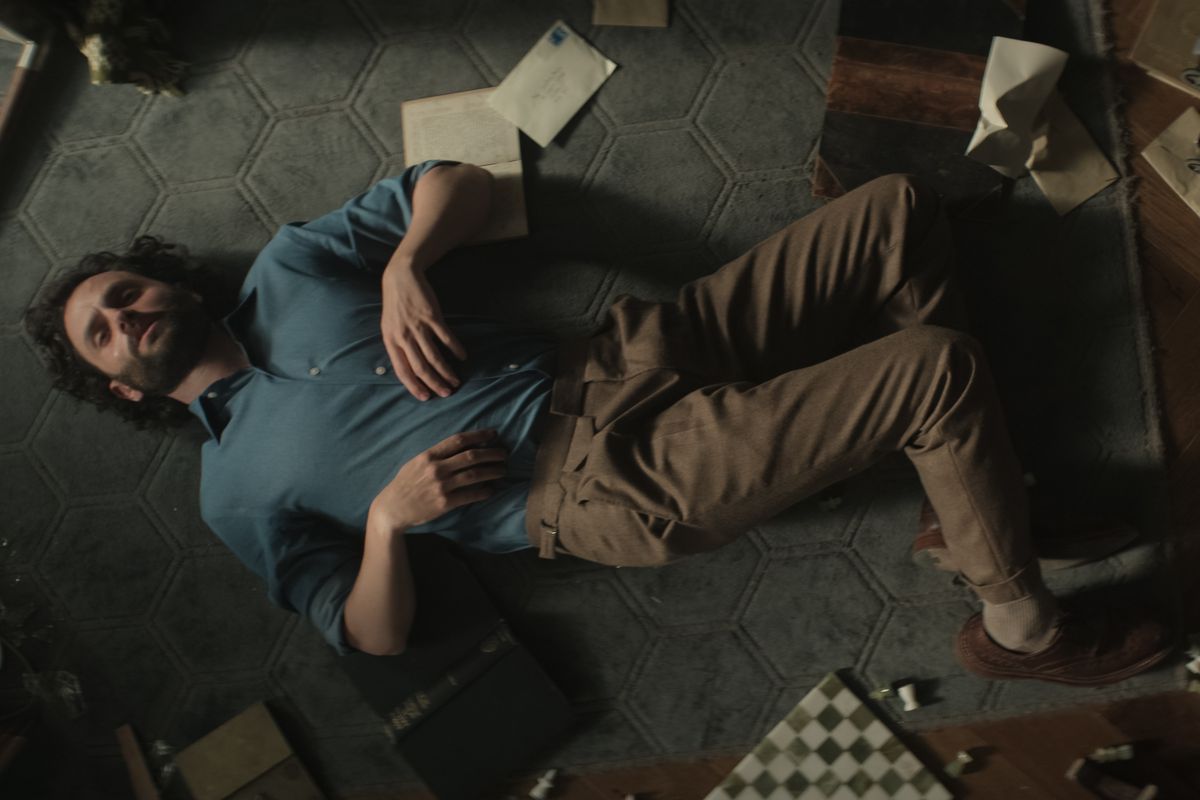 Joe Goldberg giace sul pavimento del suo appartamento con libri, carte e una scacchiera intorno a lui nella quarta stagione di You.