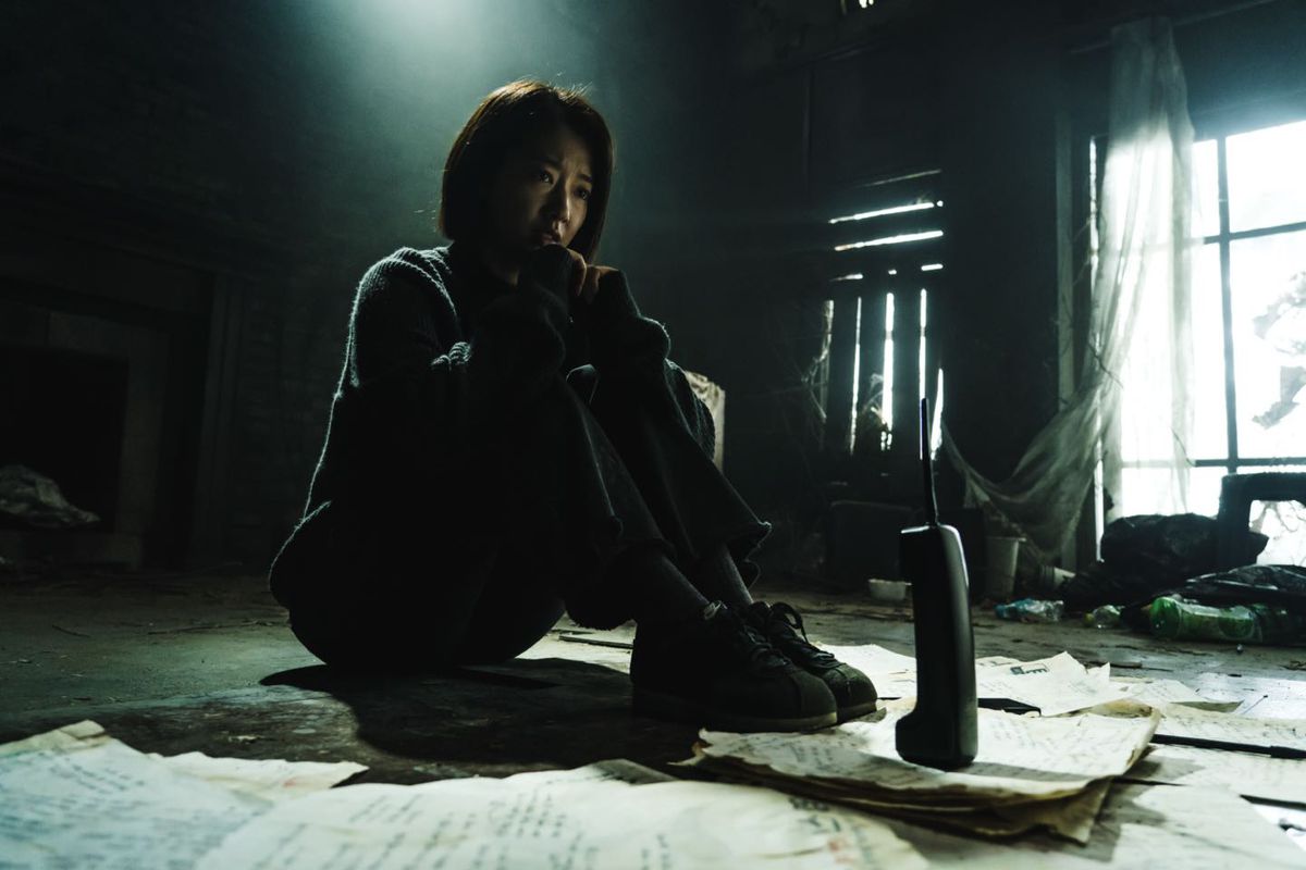 Park Shin-hye seduto di fronte a un telefono cordless in un soggiorno buio e fatiscente in The Call (2020)