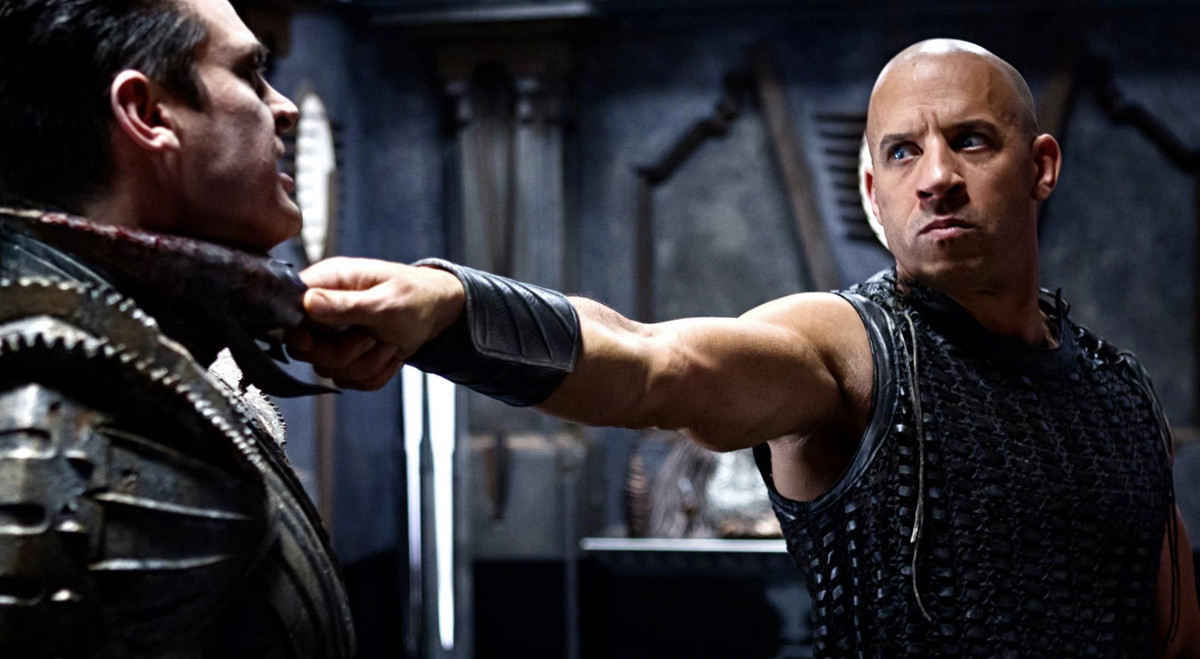 Riddick (Vin Diesel) tiene una lama frastagliata e ricoperta di sangue alla gola di un uomo con un'armatura di metallo ricoperta di lame di una sega rotante in Riddick del 2013