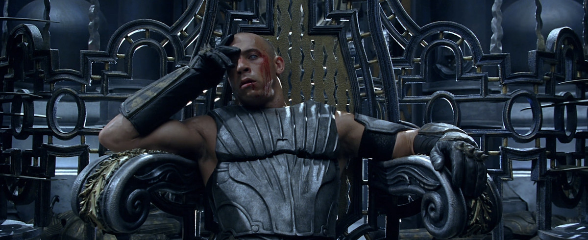 Riddick (Vin Diesel), grondante di sangue e con indosso un'armatura d'acciaio grigia, siede su un trono con la testa appoggiata sulla mano in Le cronache di Riddick