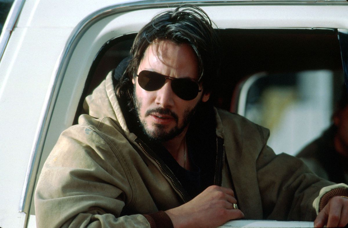 Keanu Reeves ha un bell'aspetto con occhiali da sole, capelli arruffati e una giacca marrone sul sedile del passeggero di un'auto in The Gift.