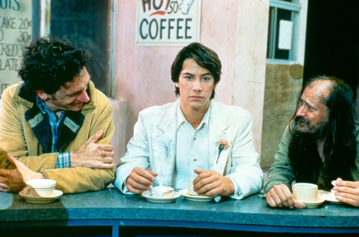 Un giovane Keanu Reeves, che indossa un abito bianco con un fiore rosa appuntato sopra, siede al bancone di un ristorante tra due uomini che gli parlano ma sembra che non stia ascoltando.