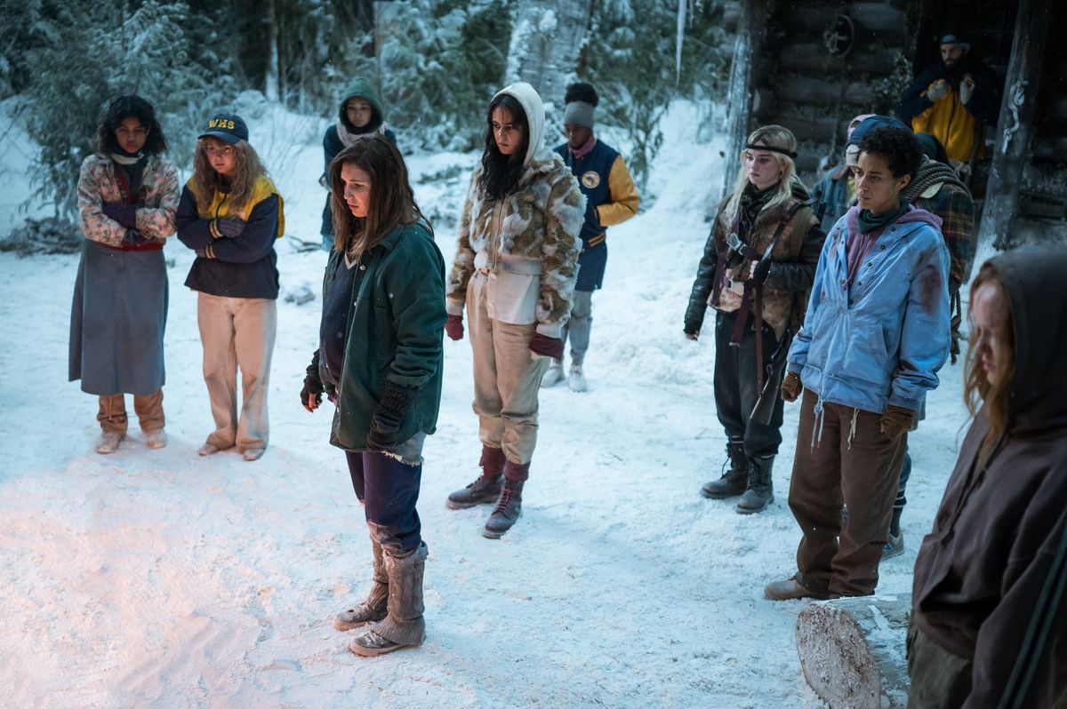 Gli adolescenti Yellowjackets stanno nella neve fuori dalla loro cabina, con un falò appena fuori dallo schermo nella premiere della seconda stagione di Yellowjackets.