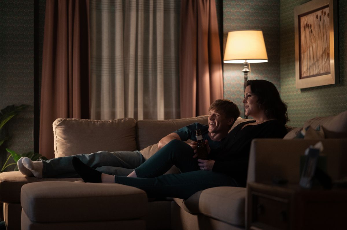 Jeff e Shauna sorridono insieme su un divano in giacche gialle.  Si sdraia, vicino all'incavo del suo braccio mentre lei si siede.
