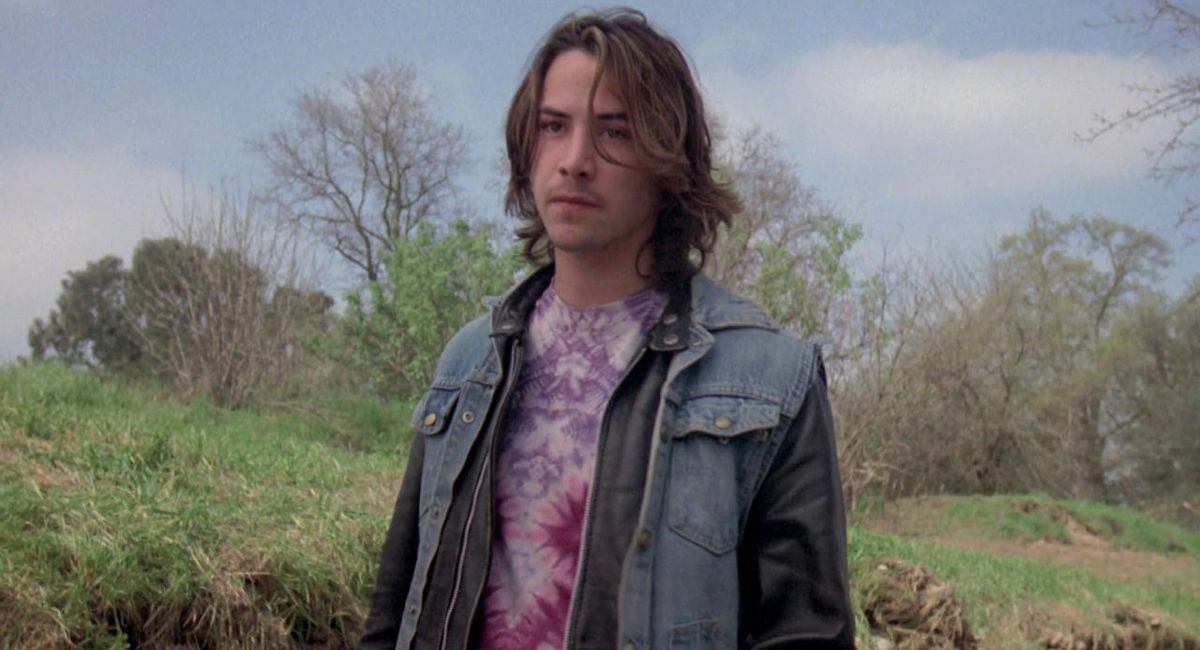Un giovane Keanu Reeves con i capelli lunghi indossa una giacca di jeans e una camicia tie-die su uno sfondo di erba e alberi in River's Edge.