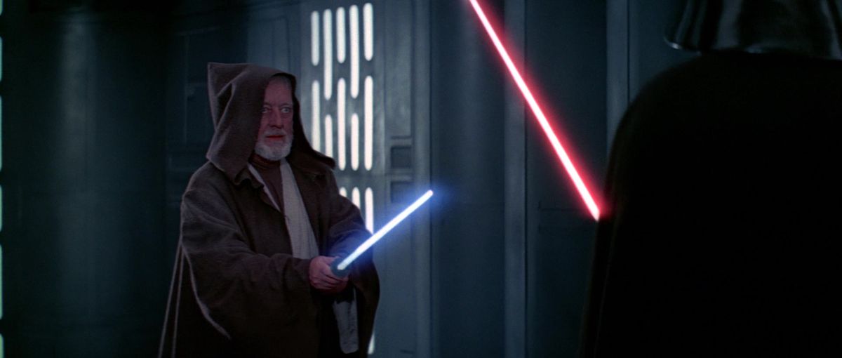 1977 Obi-Wan Kenobi (Alec Guinness), in abiti Jedi e con il cappuccio a punta tirato su per nascondere parzialmente il volto, solleva la sua spada laser mentre Darth Vader incombe sul bordo dell'immagine in Star Wars: Una nuova speranza