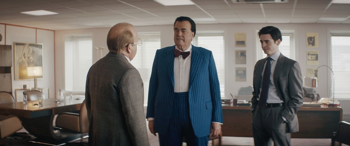 Tre uomini in giacca e cravatta degli anni '80 si trovano in un ufficio.  Uno di loro è molto alto e grosso e indossa un papillon.