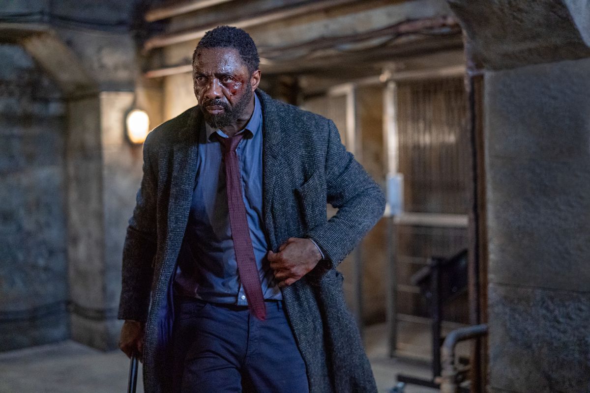 Un uomo (Idris Elba) vestito con una camicia elegante blu, cravatta bordeaux e un lungo cappotto di tweed con lividi visibili e sangue sul viso.