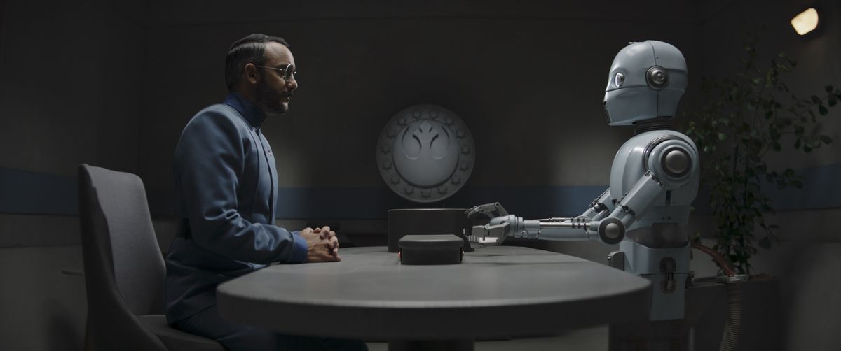 Il dottor Pershing siede di fronte a un robot bianco e sottile a un tavolo grigio in The Mandalorian