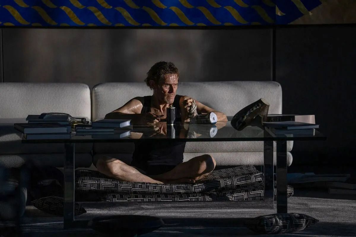 Un uomo nudo e spettinato in boxer (Willem Dafoe) siede a gambe incrociate davanti a un tavolo di vetro in un soggiorno buio con la luce che entra da una finestra fuori campo in Inside.