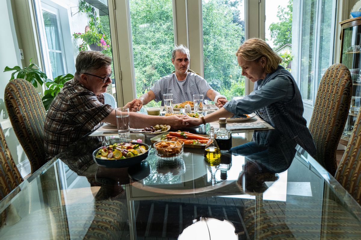 Nathan Lane, Joaquin Phoenix e Amy Ryan siedono a tavola tenendosi per mano e pregando in Beau is Afraid
