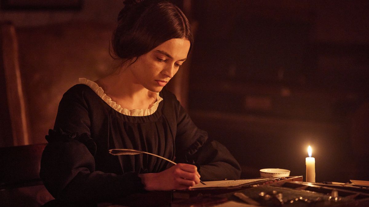 Emma Mackey nei panni di Emily Brontë che scrive su un pezzo di pergamena con una penna d'oca a lume di candela in Emily.