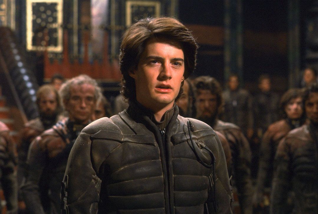 Un giovane (Kyle Maclachlan) con i capelli castani vestito con un polveroso abito high-tech si trova in un atrio circondato da persone vestite con abiti simili in Dune (1984).