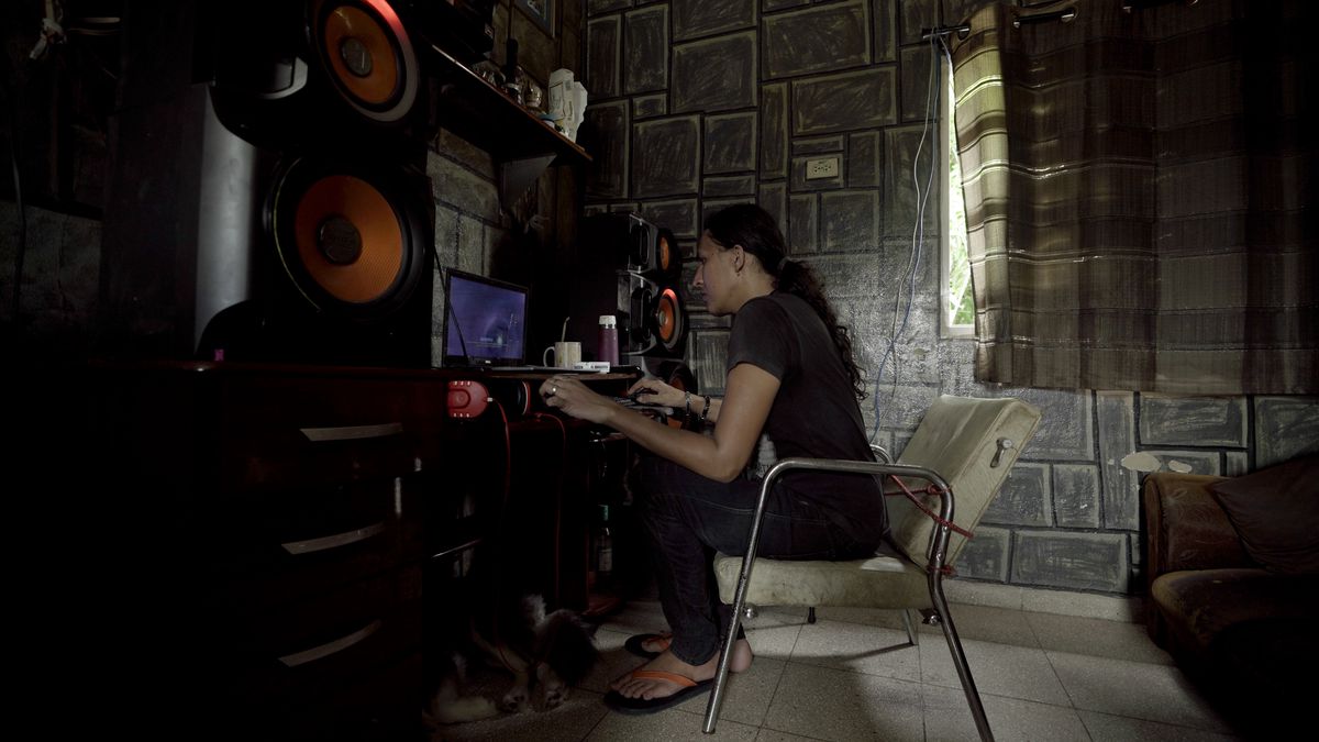 Un giovane con i capelli neri legati sulla schiena siede su una sedia bianca in una stanza con muri di pietra scarsamente illuminata a Cuba mentre gioca a un videogioco su un piccolo computer portatile