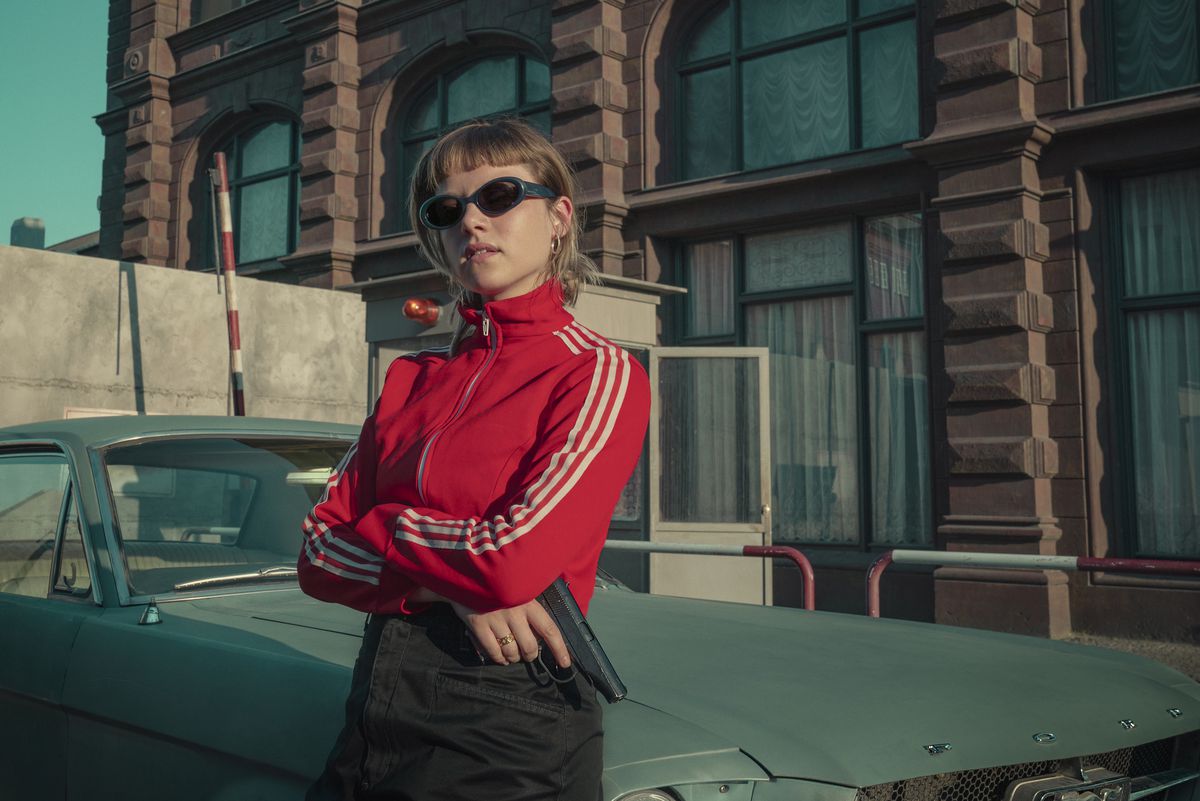 Jelia Haase nei panni di Kleo indossa una tuta rossa e jeans e tiene in mano una pistola mentre si appoggia a un'auto.  Indossa anche occhiali da sole e ha un bell'aspetto.