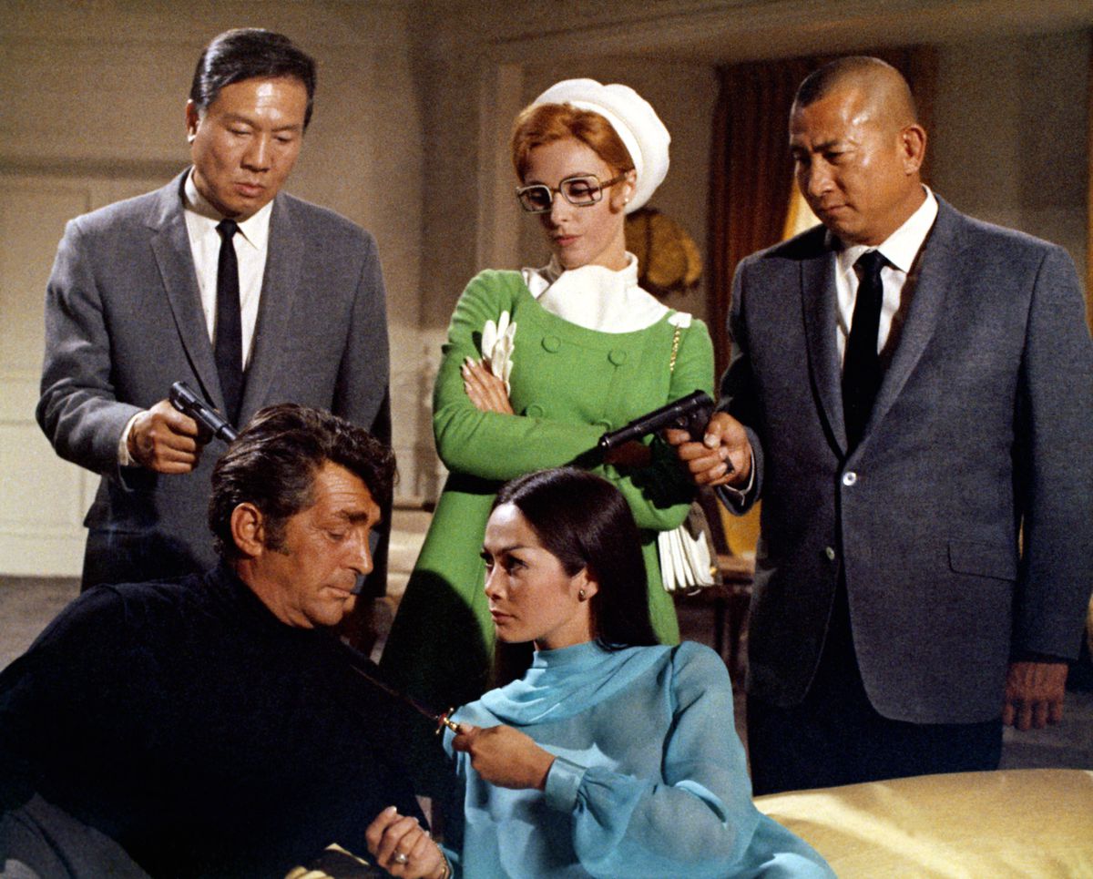 Due scagnozzi asiatici puntano le pistole contro Matt Helm mentre una donna dai capelli rossi con gli occhiali e un cappotto verde sogghigna dietro di lui e una donna asiatica con un abito blu alza un coltello