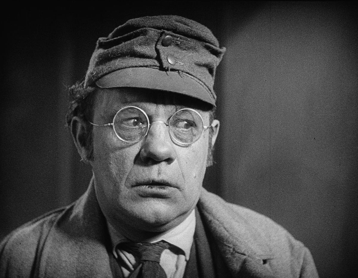Un uomo che indossa occhiali rotondi e un cappello militare osserva qualcosa fuori dallo schermo in Spione