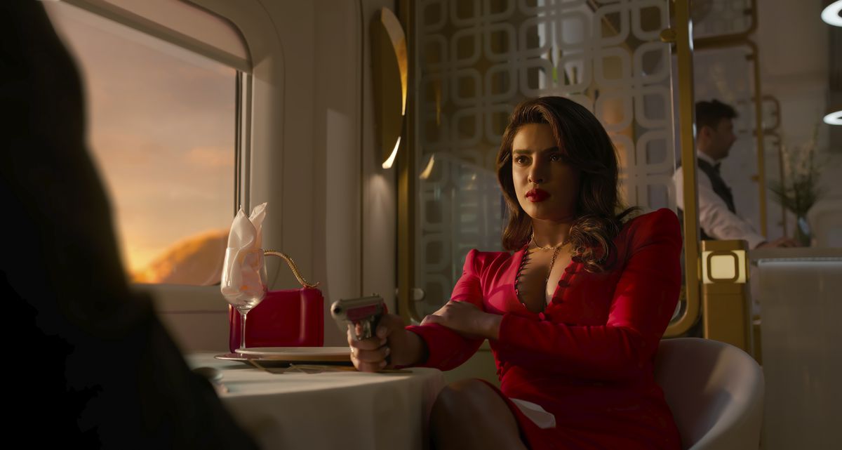 Priyanka Chopra Jonas nei panni di Nadia Sinh siede a un tavolo da pranzo su un treno mentre il sole tramonta dal finestrino, illuminando il suo vestito rosso e la pistola che punta contro un uomo fuori dallo schermo di fronte a lei nello spettacolo Prime Video Citadel.