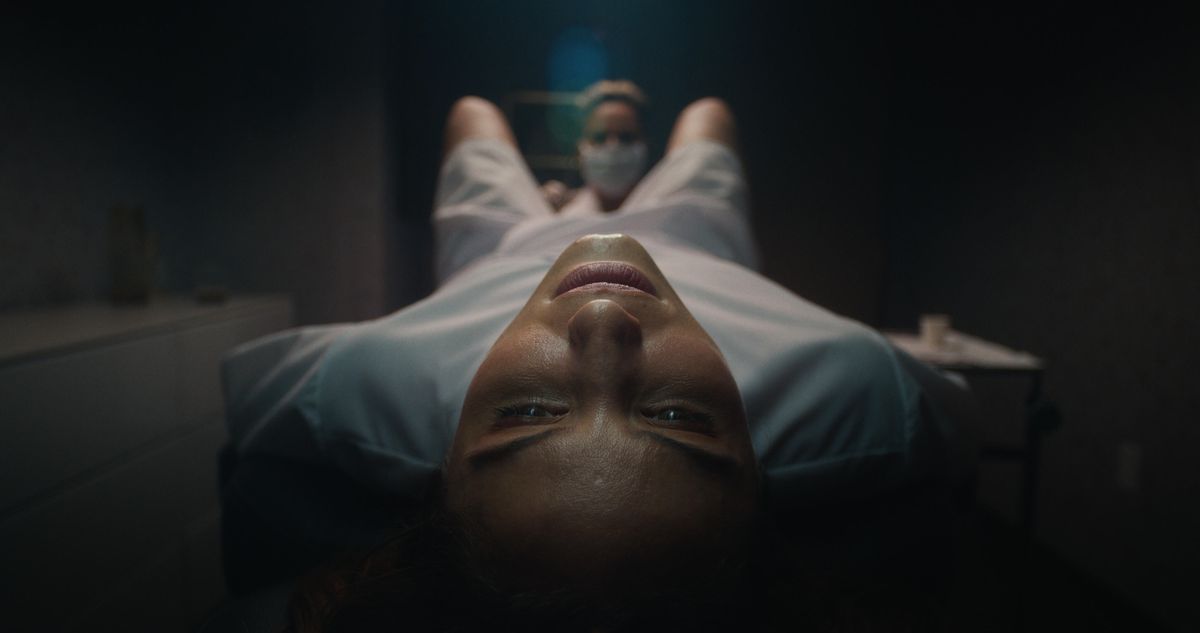 Un'inquadratura ravvicinata di una donna (Dianna Agron) in camice da ospedale sdraiata su un tavolo mentre un'infermiera le esamina tra le gambe in Clock.