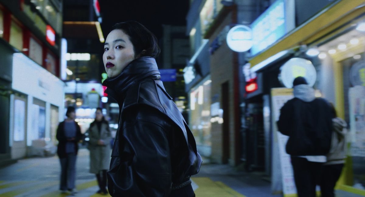 Jin-Min Park nei panni di Freddie, che indossa un rossetto rosso scuro in un cappotto di pelle nera in piedi nel mezzo di una strada di una città coreana di notte in Return to Seoul.