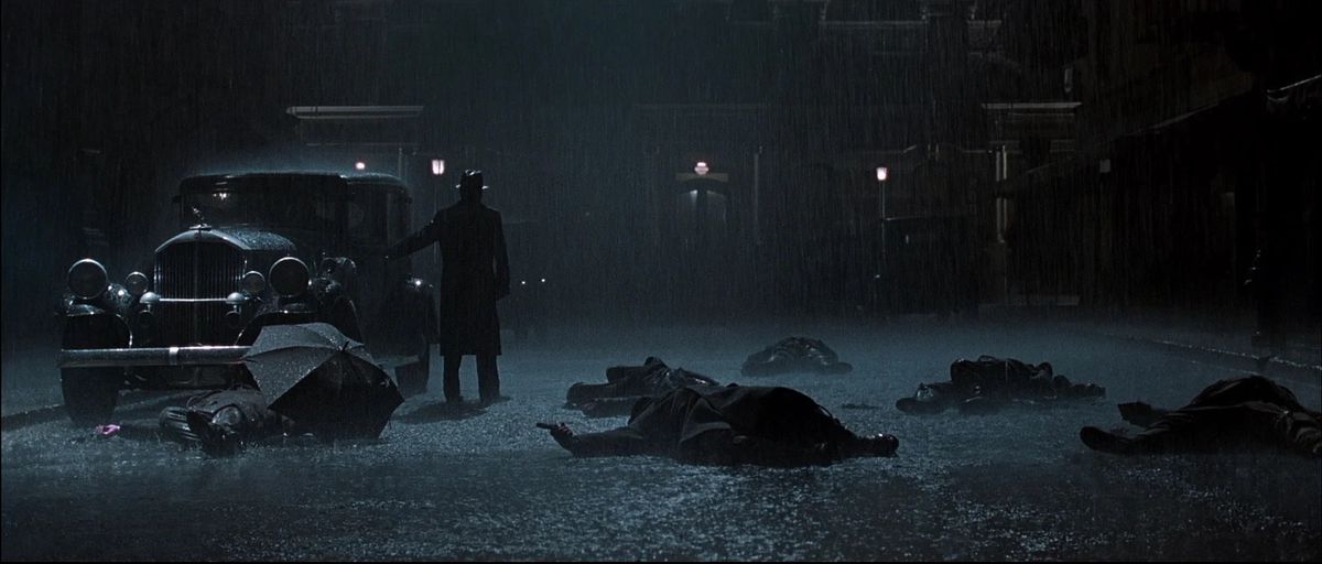 Un uomo con cappello e impermeabile si trova in mezzo a una strada buia accanto a un'automobile circondata da cadaveri mentre piove a dirotto.